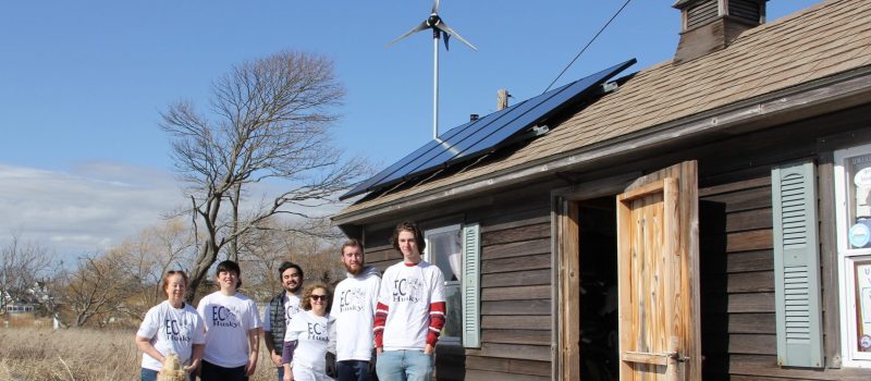 UConn Avery Point EcoHusky Club members and Syma Ebbin, 3rd from left, faculty advisor and CTSG research coordinator, gather at a campus building with solar panels and a small wind turbine installed thanks to the club’s work.