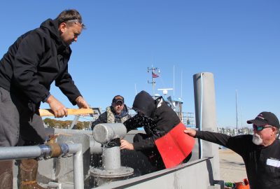 Students practice fixing a leak while another participate hands them a piece of neoprene to add to the repair.